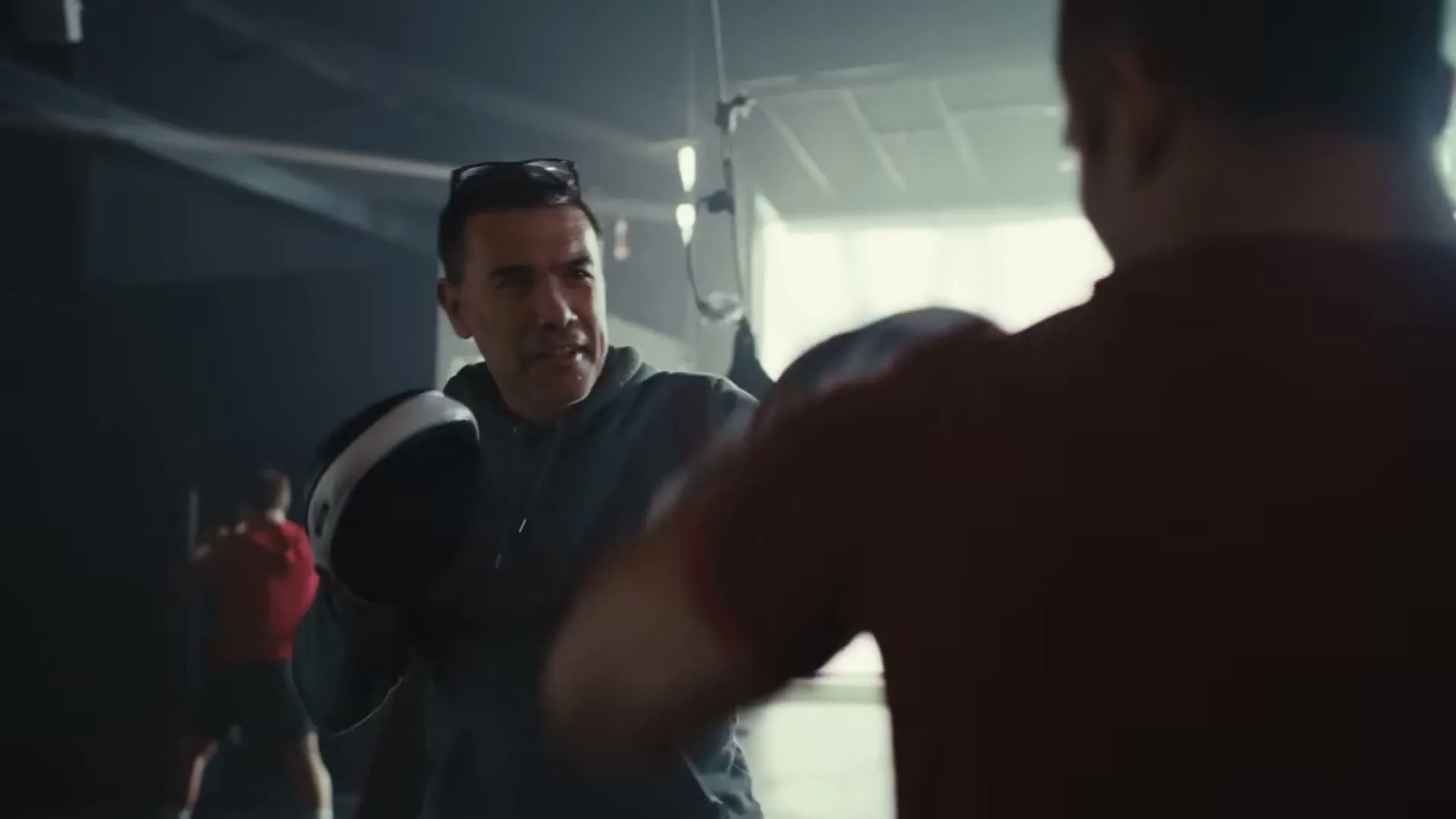 A man with Down syndrome in a grey hoodie and glasses on his head practices boxing with a trainer in a dimly lit gym.