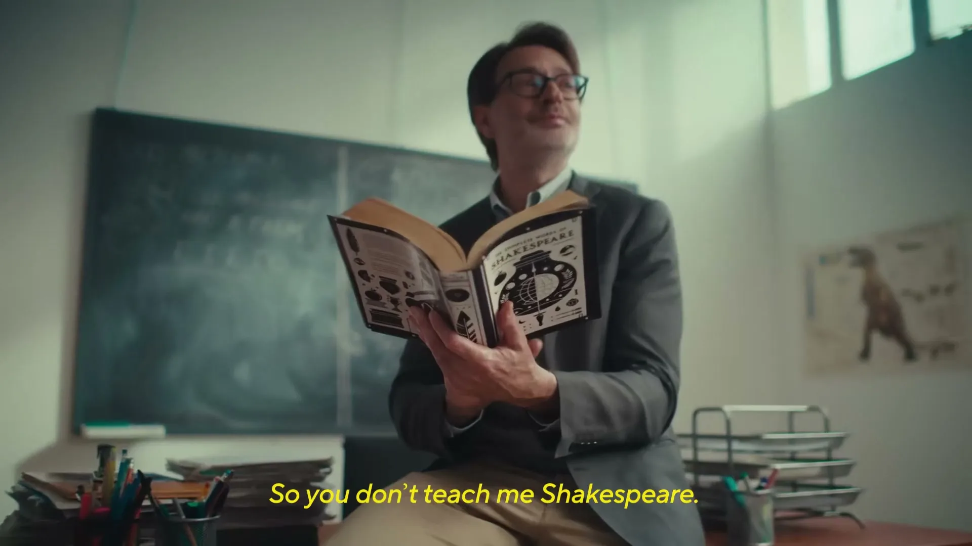 A male teacher in his 40s, wearing a grey suit and glasses, sits on a desk in a classroom, holding an open book and looking up with a thoughtful expression.
