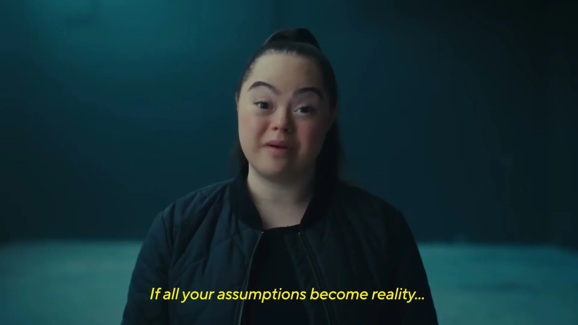 A young woman with Down syndrome, wearing a black bomber jacket, speaks directly to the camera in a dimly lit studio.