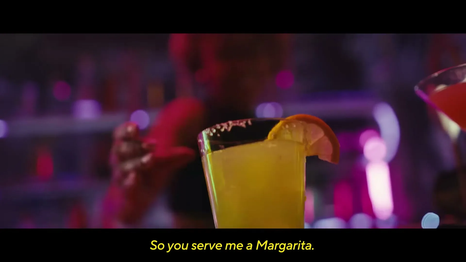 A woman with Down syndrome smiles as she serves a margarita at a bar, with colorful bottles and neon lights in the background.