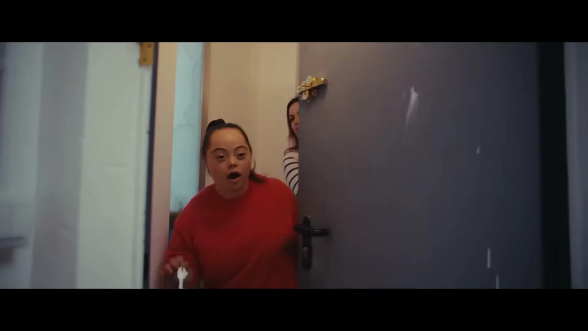 A young woman with Down syndrome in a red top bursts through a doorway, looking surprised, followed by another woman in a striped top.