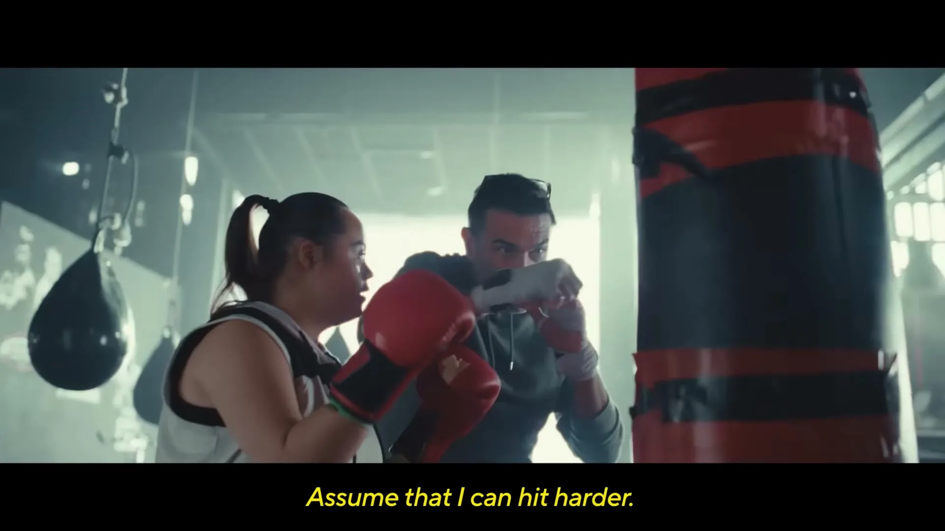 A young woman with Down syndrome practices boxing with a male trainer in a gym, hitting a red punching bag.
