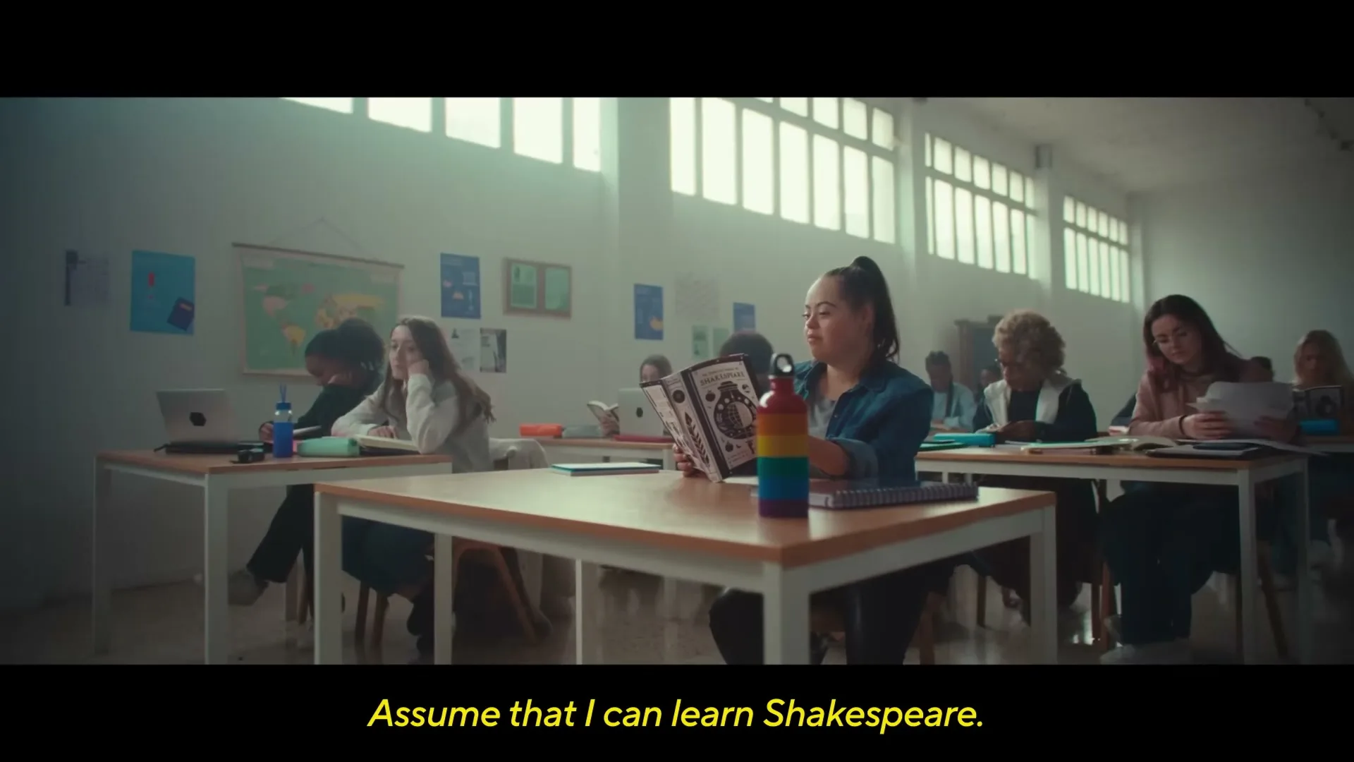 A young woman with Down syndrome sits at a desk in a classroom, reading a book, surrounded by other students.