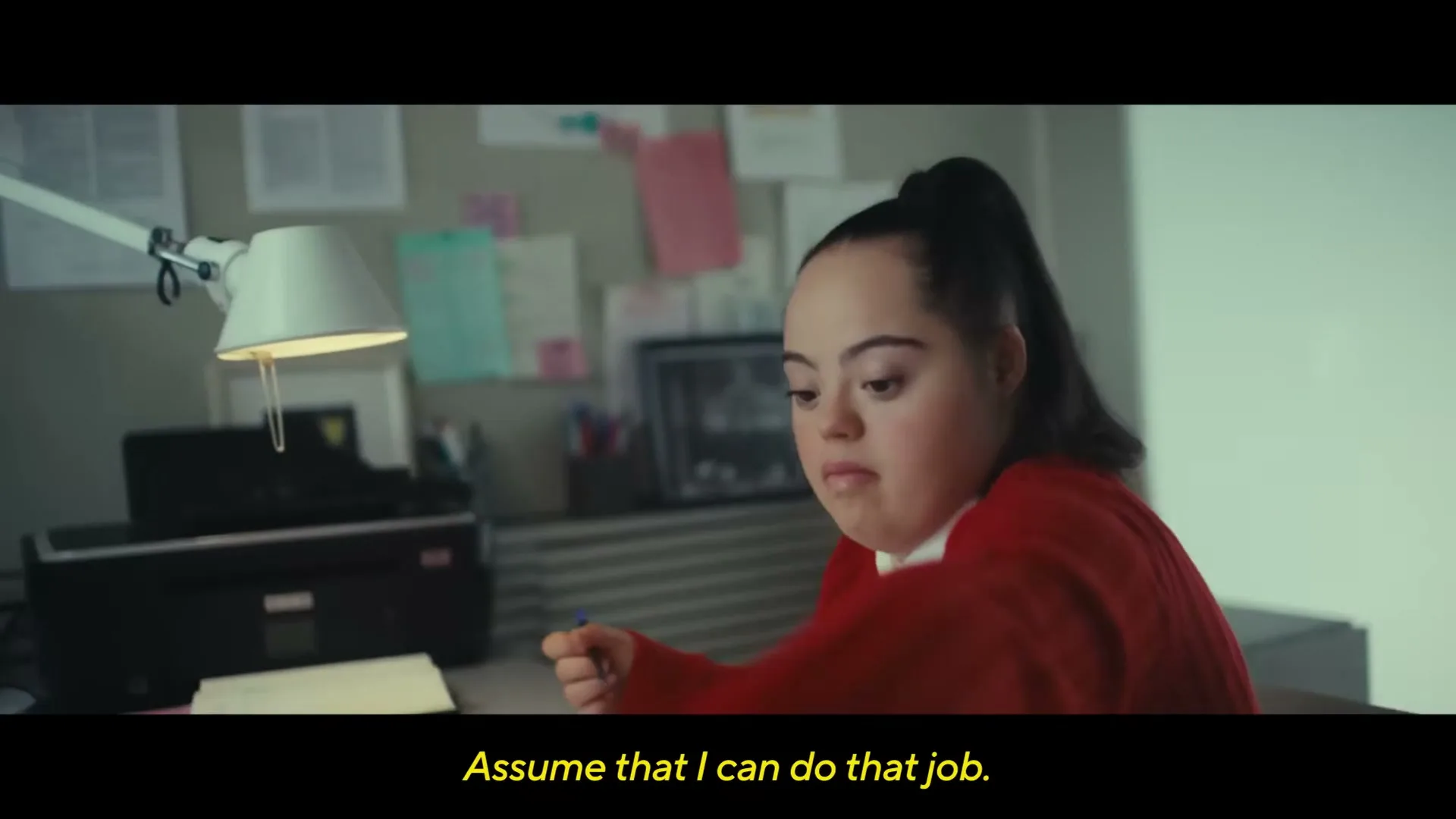 A young woman with Down syndrome, wearing a red sweater, sits at an office desk, writing on papers with a pen.
