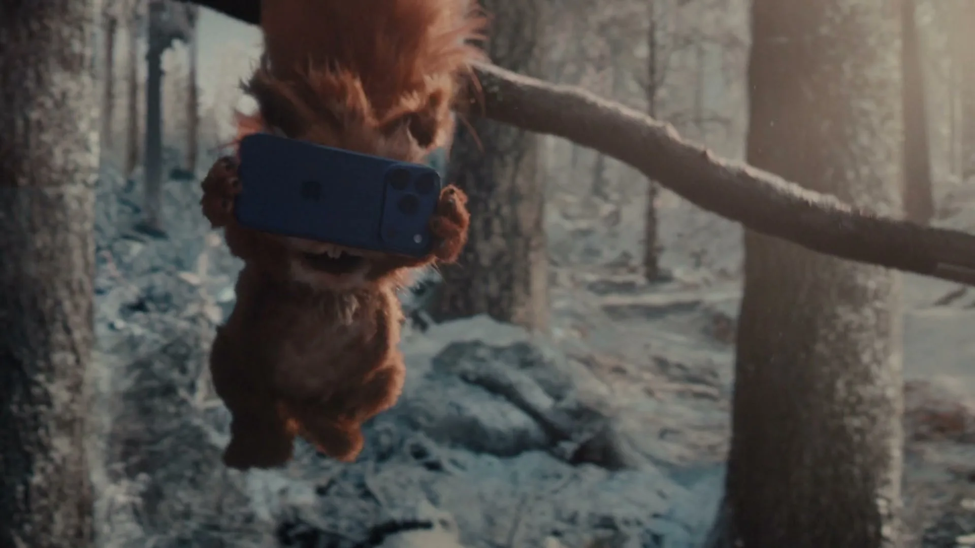 A cute, fluffy, animated squirrel hangs upside down from a tree branch, holding a blue iPhone in its paws in a snowy forest.