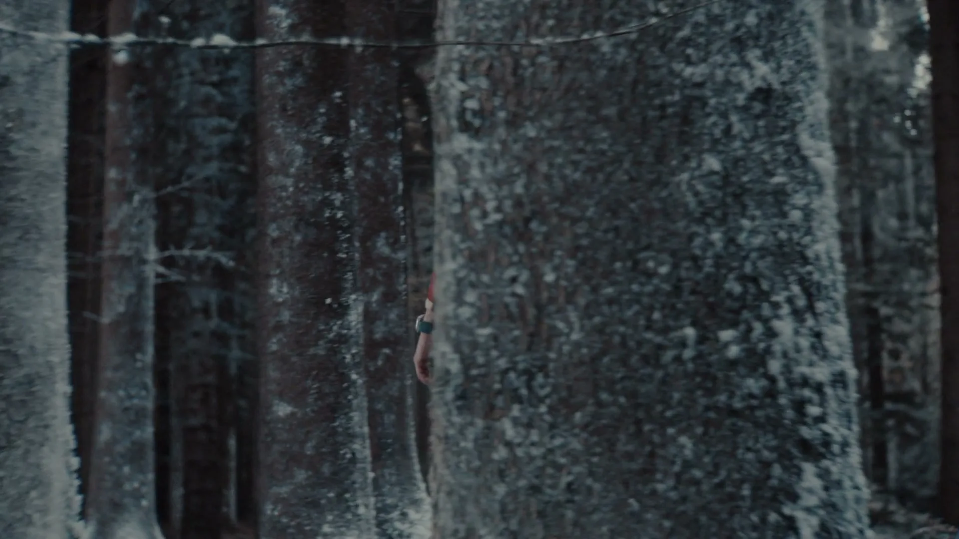 A man in a red jacket and backpack walks through a snowy forest, checking his smartwatch as he goes.