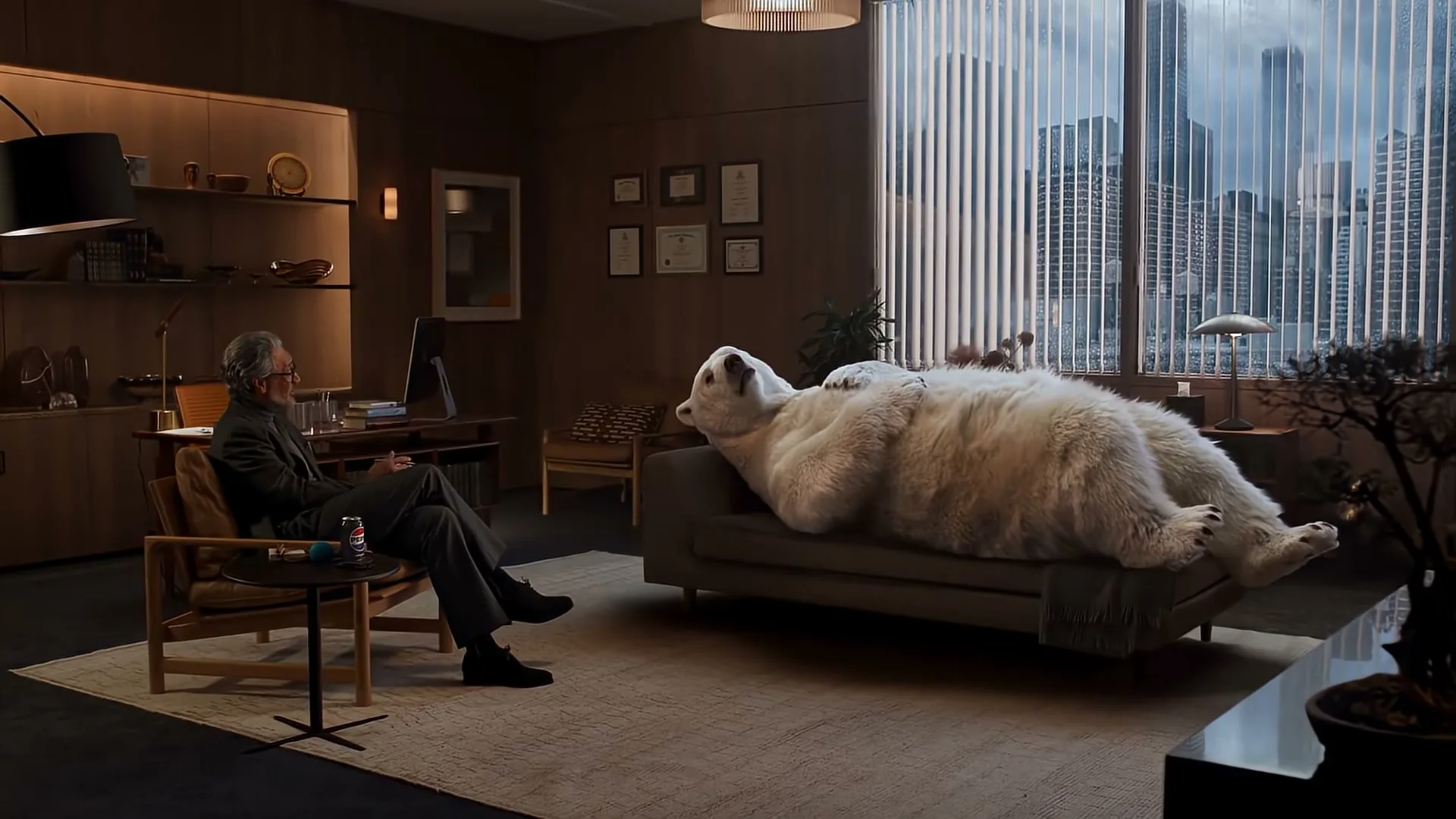 A man in a suit sits in a chair, looking at a large polar bear lying on a couch in a modern office with a city view.