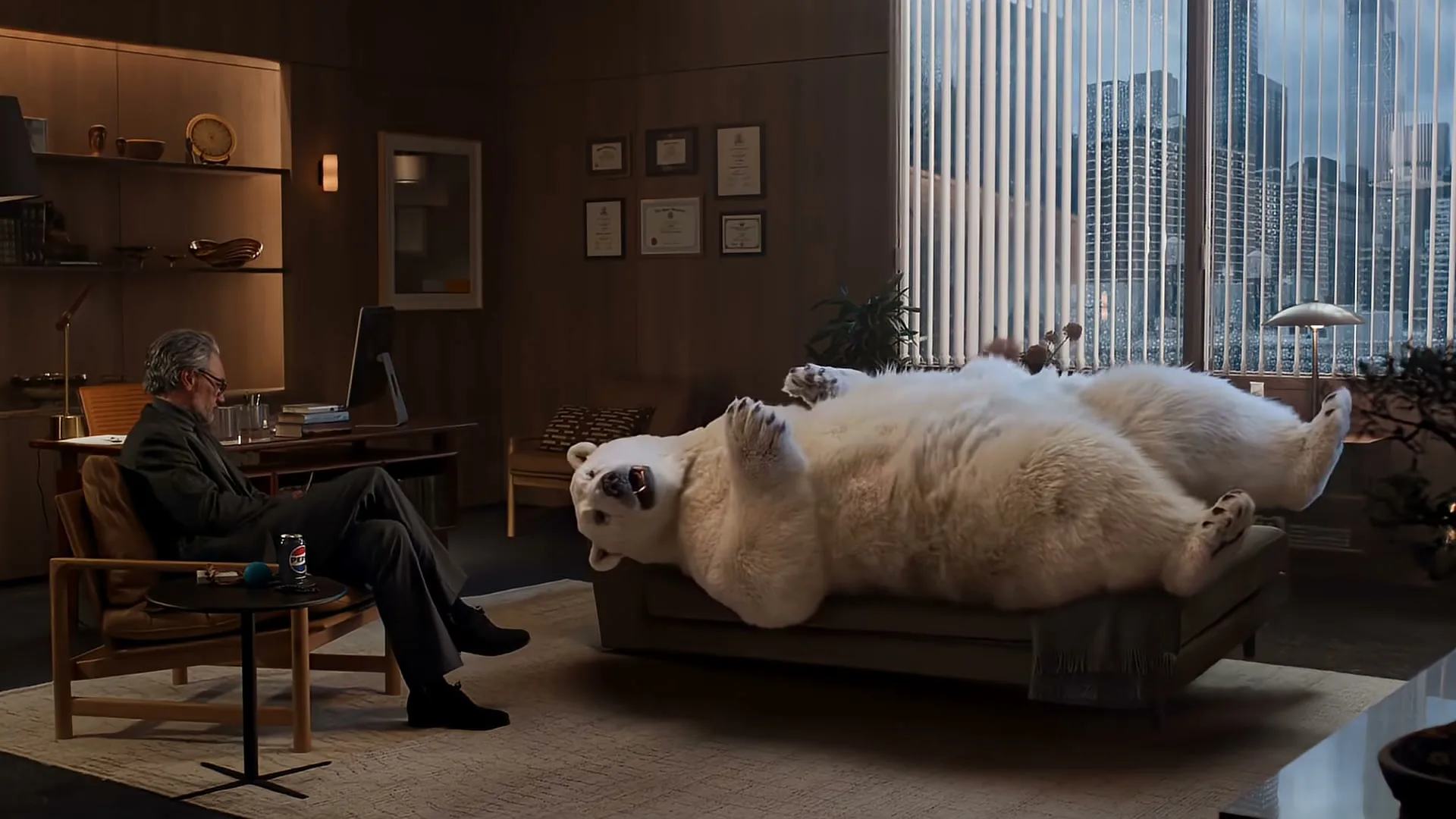 A man in a suit sits in a chair, looking at a large polar bear lying on a couch in a modern office with a city view.