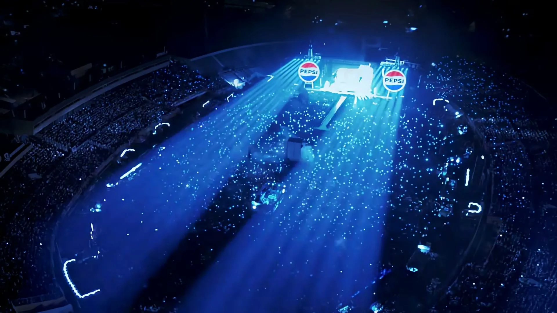 An aerial view of a stadium at night, filled with a crowd holding glowing blue lights, with two large Pepsi logos on screens at the far end.