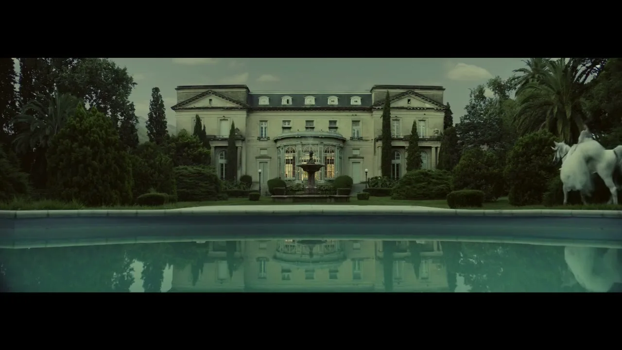 A wide shot of a grand mansion with a swimming pool in the foreground, reflecting the building, as a white horse walks by on the right.