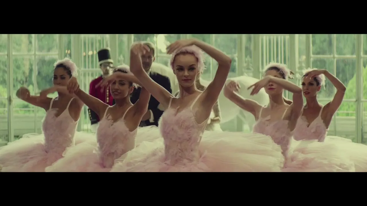 Five ballerinas in pink tutus perform a synchronized dance in a grand, glass-walled room, while a man in a suit and another in a red uniform watch in the background.