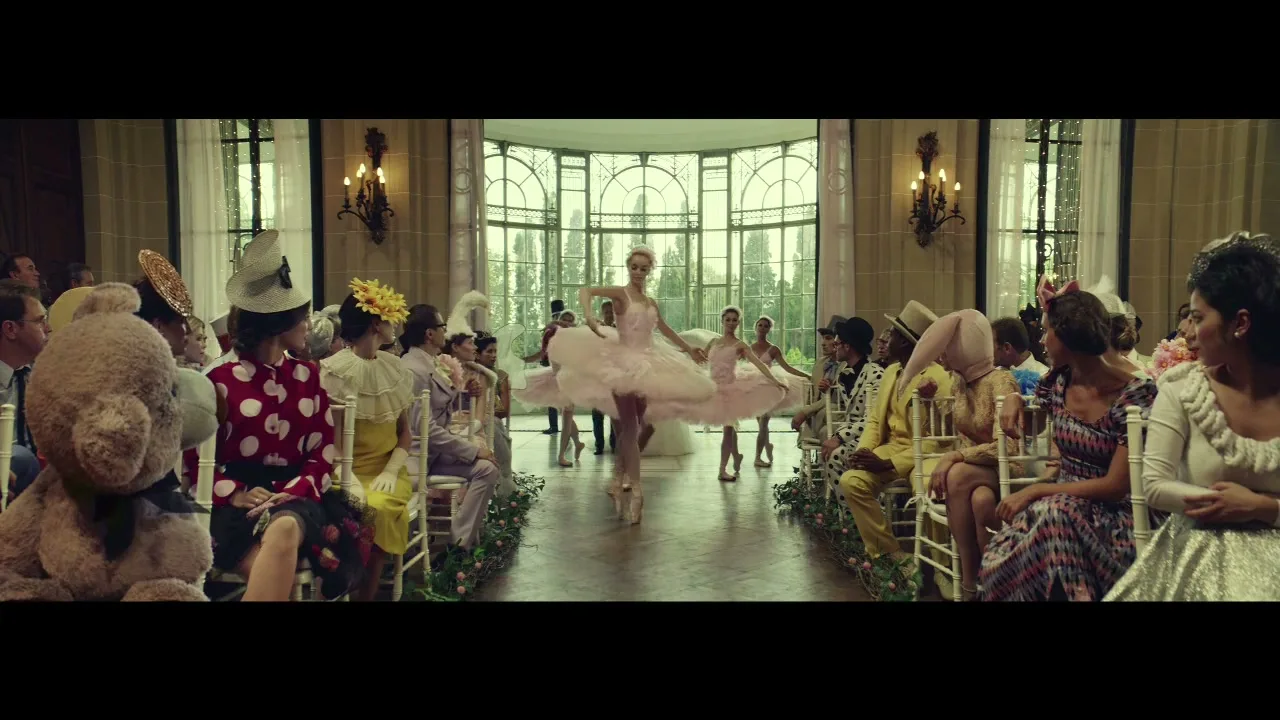 A group of people in eccentric outfits sit on chairs, watching a ballerina in a pink tutu dance down an aisle in a grand hall.