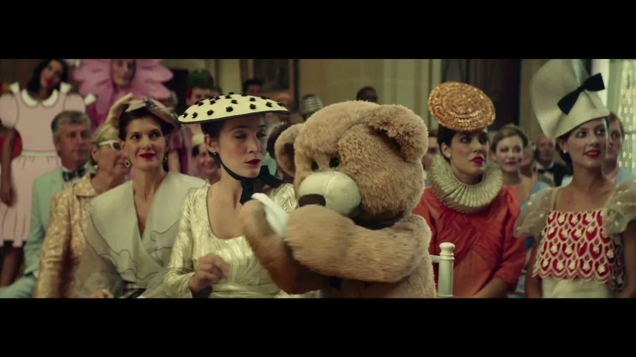 A woman in a polka dot hat and a teddy bear in a bow tie sit in a church pew, surrounded by other guests in elaborate hats.