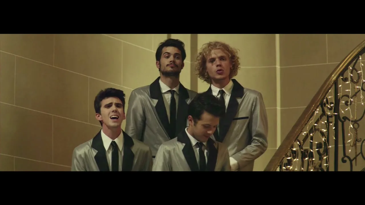 Four young men in matching silver and black tuxedos sing on a grand staircase, looking up and performing in a formal interior setting.