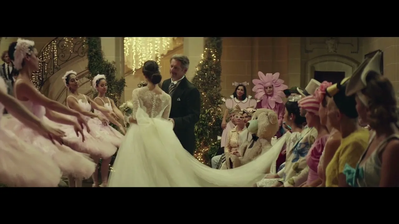 A bride in a white gown and a groom in a tuxedo stand at the altar, surrounded by ballerinas and guests in whimsical costumes at a wedding.