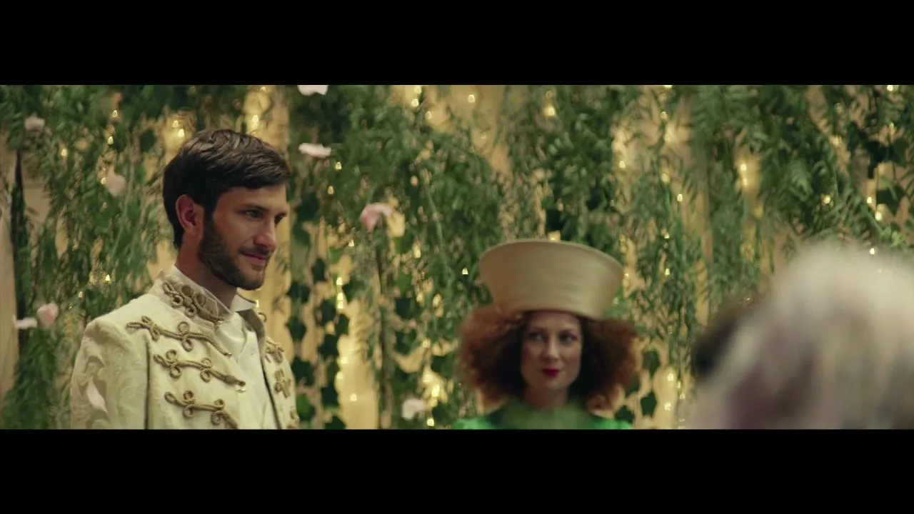 A groom in an ornate white jacket smiles, standing next to a woman in a green dress and unique hat, at a wedding ceremony.
