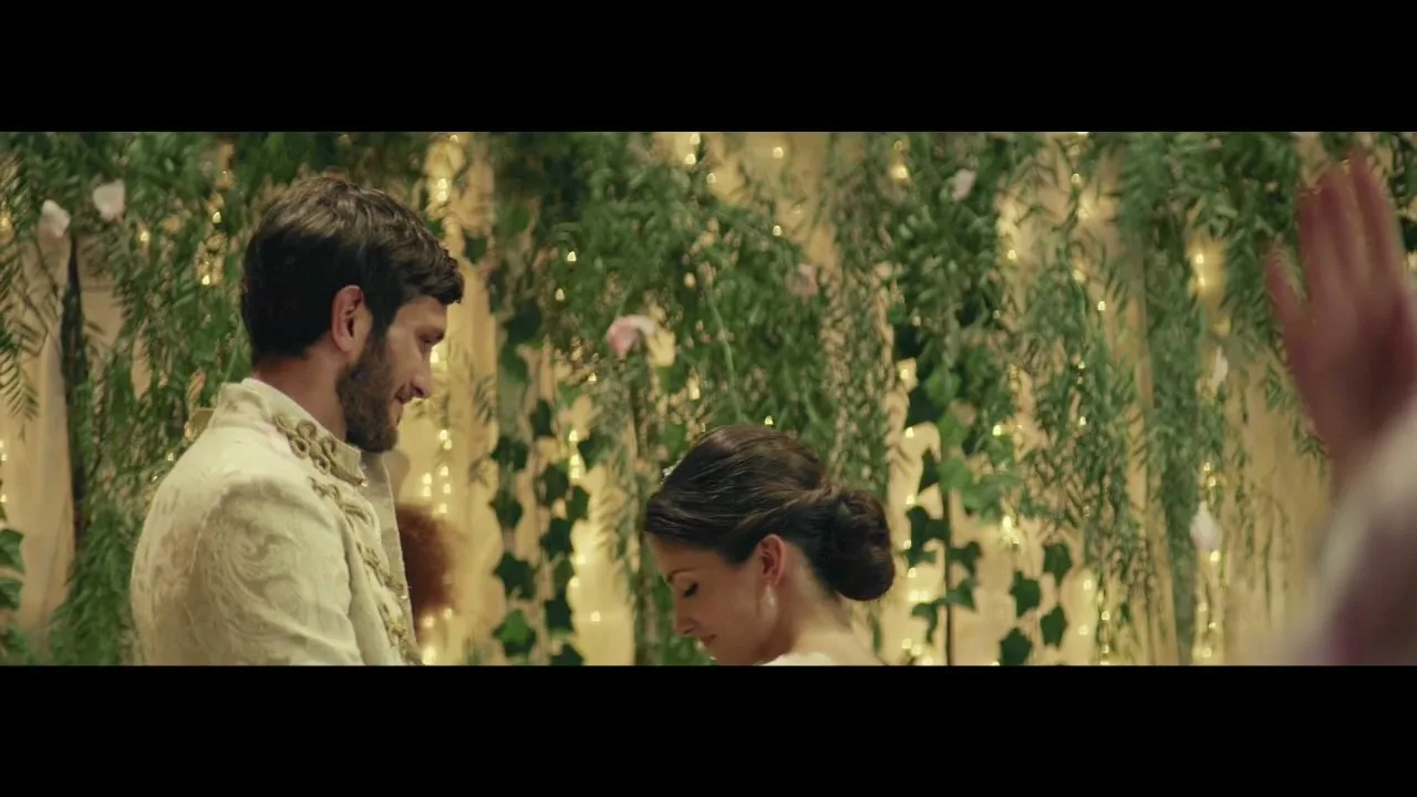 A groom in an ornate white suit looks at his bride, who is wearing a white dress and tiara, as they stand in front of a wall of greenery and fairy lights.