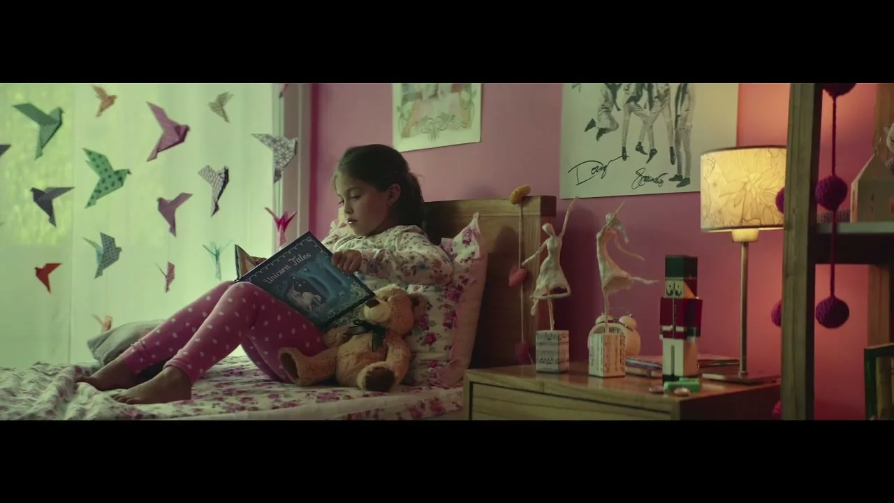 A young girl in pajamas sits on her bed, reading a book titled "Unicorn Tales" with a teddy bear beside her in a pink bedroom.