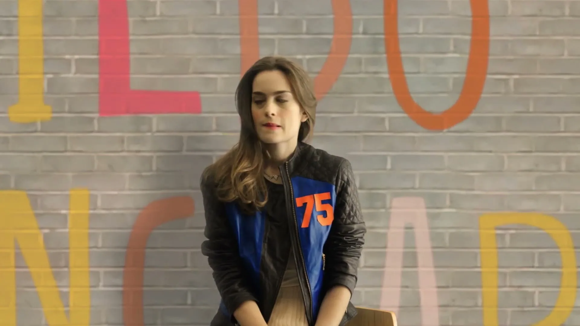 A young woman with long brown hair, wearing a black and blue jacket with "75" on it, sits in front of a brick wall with colorful letters.