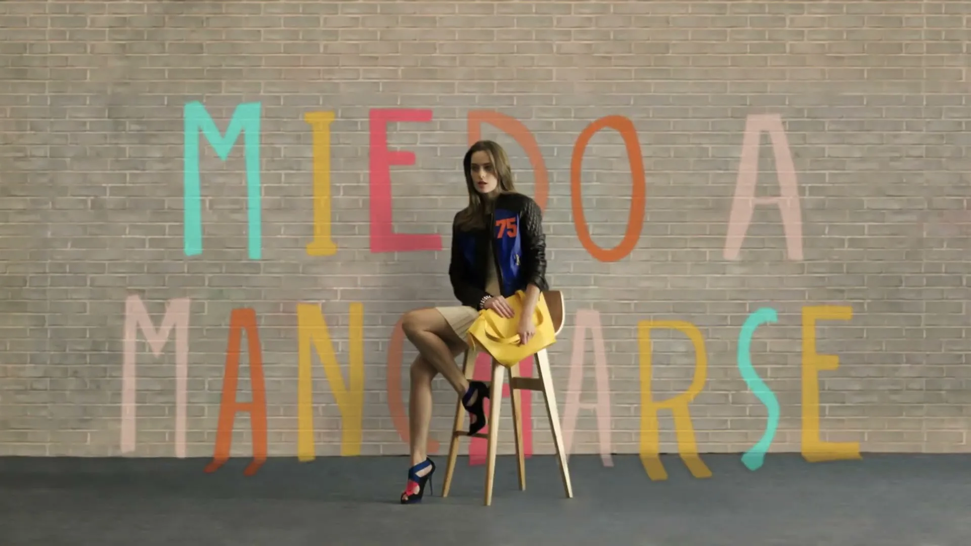 A young woman with long brown hair, wearing a black and blue jacket, yellow skirt, and colorful heels, sits on a stool in front of a brick wall with colorful text.