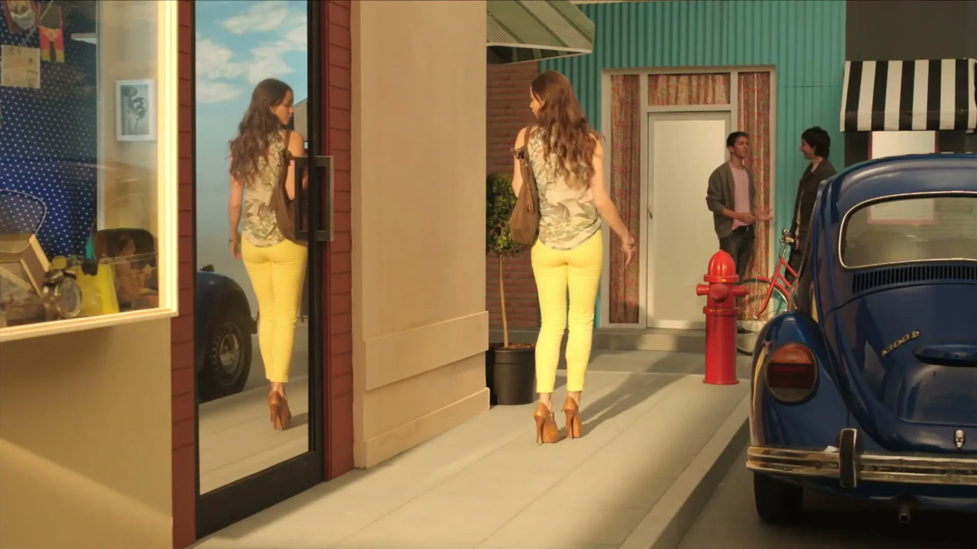 A young woman with long brown hair, wearing a patterned top and yellow pants, walks away from the camera on a stylized street set, passing a blue car and two men.
