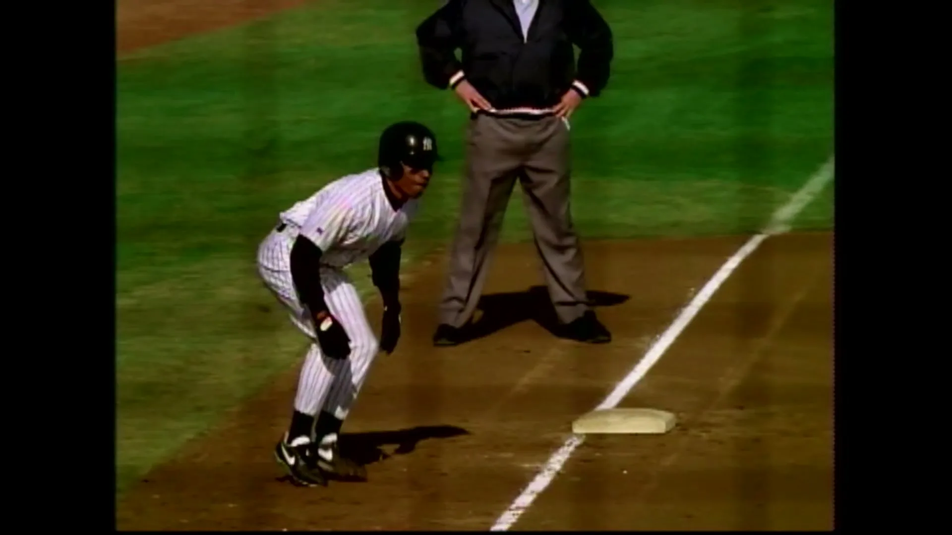 A baseball player in a white pinstripe uniform and black helmet crouches near a base, while an umpire stands behind him on a green baseball field.