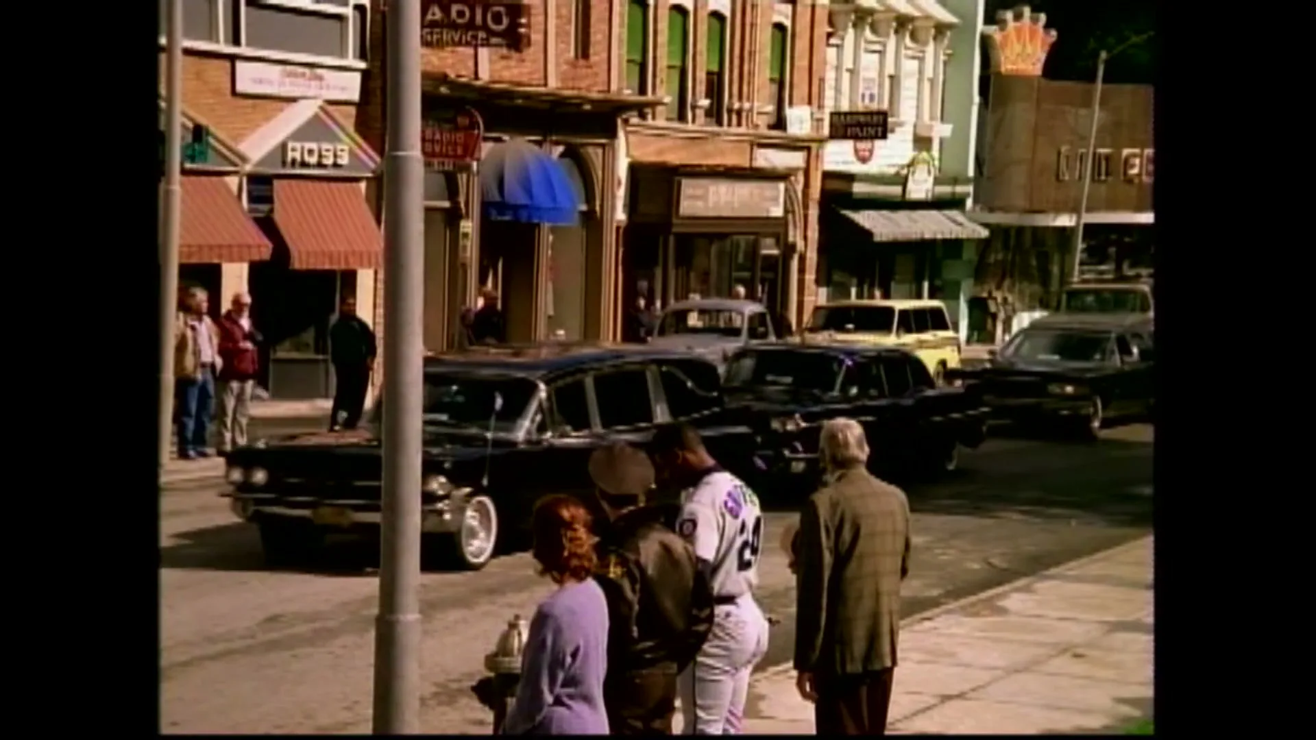 A group of people stand on a sidewalk, watching a black car drive by on a sunny day in a small town.