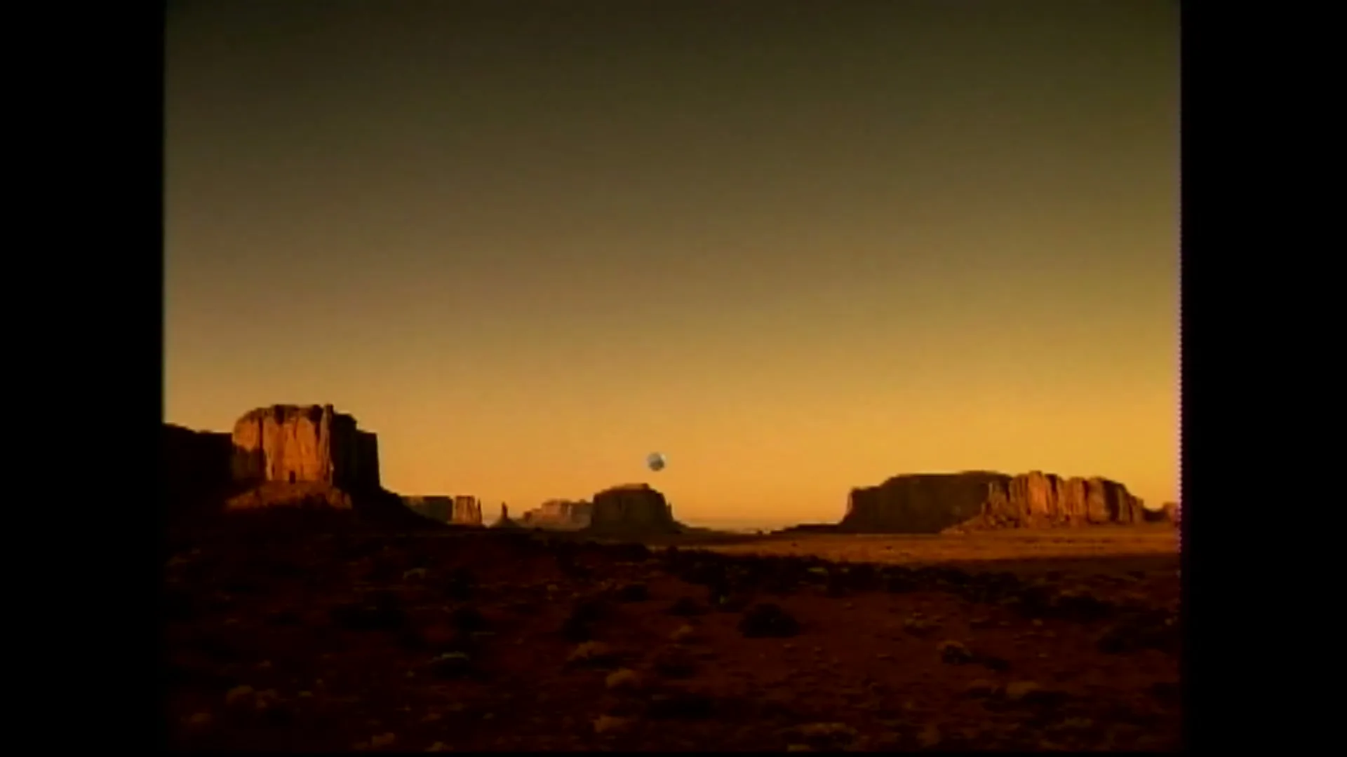 A wide shot of a desert landscape with large rock formations under a golden sky, as a basketball flies through the air.