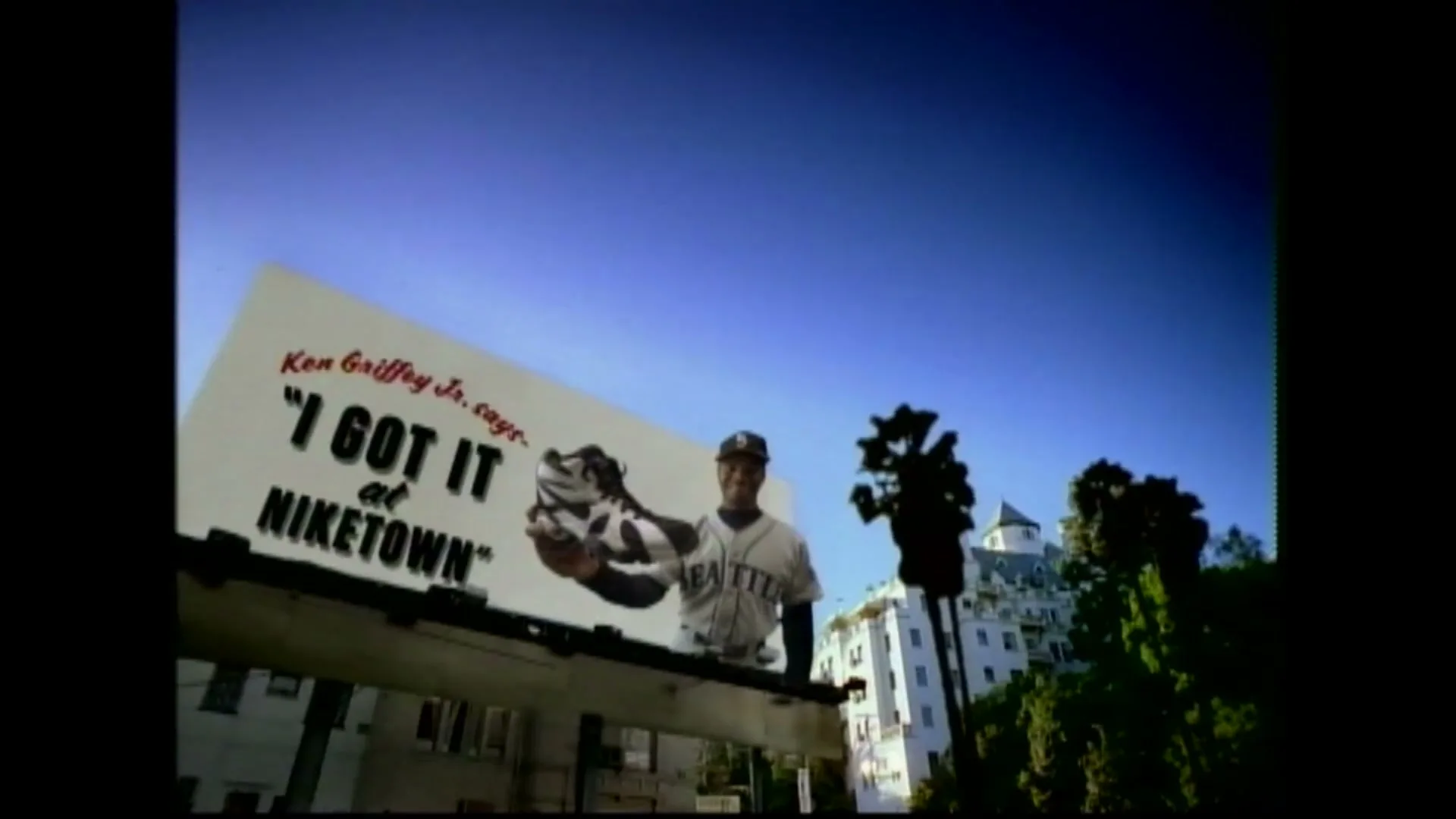 A billboard featuring baseball player Ken Griffey Jr. and a Nike shoe stands against a clear blue sky, with buildings and palm trees visible below.