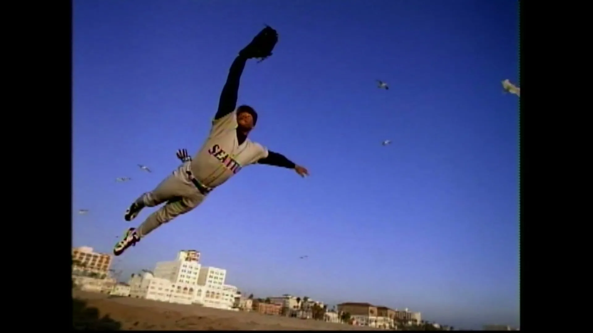 A male baseball player in a Seattle Mariners uniform dives through the air with his glove outstretched, against a clear blue sky with buildings in the background.