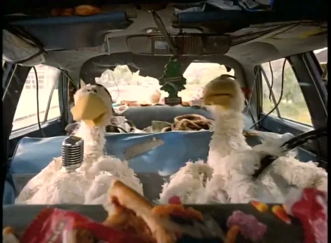 Two white chicken puppets are sitting in the front seats of a messy, old car, with one speaking into a microphone.