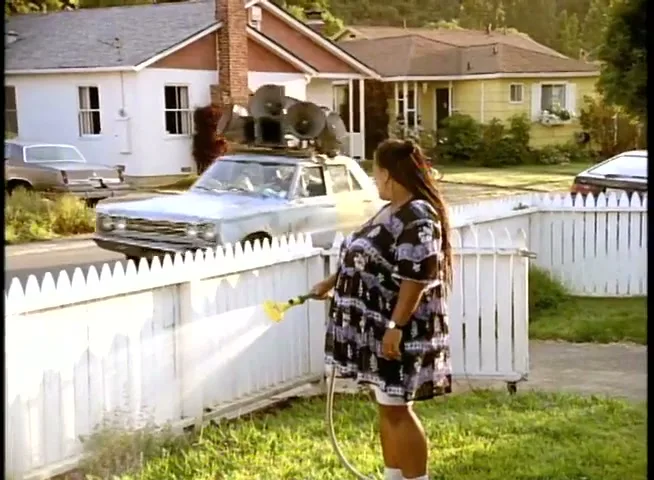 A woman with long braids and a patterned dress waters her lawn as a car with a bullhorn on its roof drives by in a suburban neighborhood.