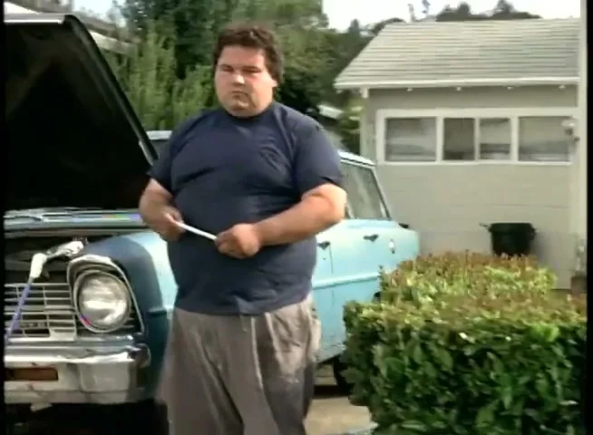 A heavy-set man in a blue t-shirt and grey pants stands in a suburban driveway, holding a tool, next to a light blue car with its hood open.
