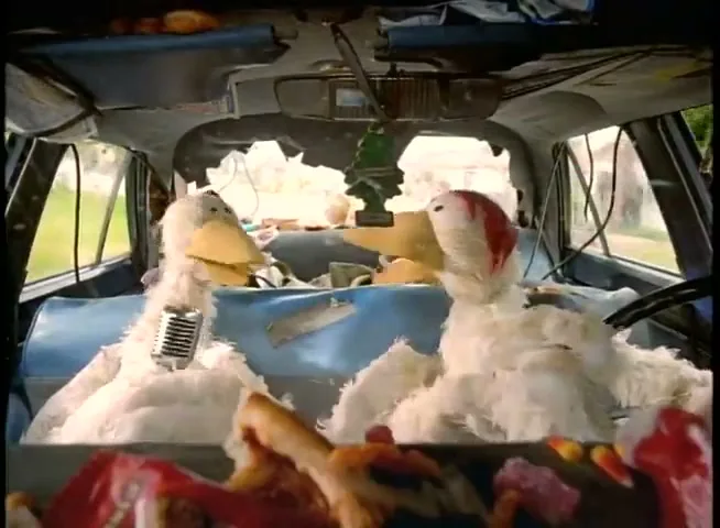 Two white duck puppets are sitting in the front seats of a messy car, with one duck speaking into a microphone.