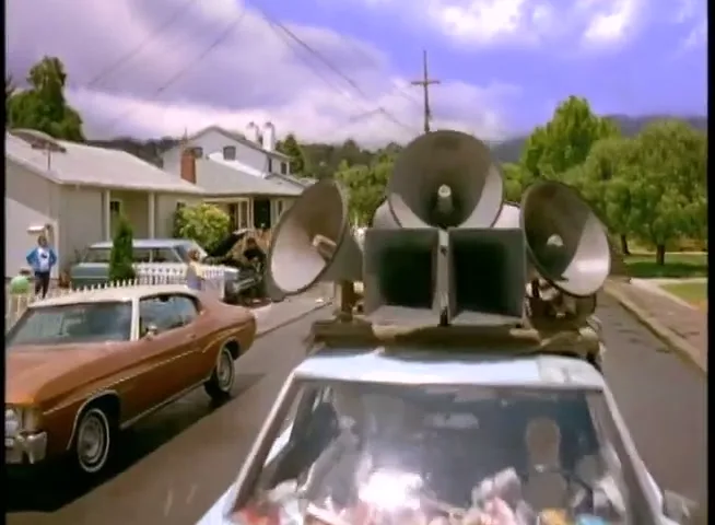 A white car with large bullhorns mounted on its roof drives down a suburban street, passing a brown car and people on the sidewalk.
