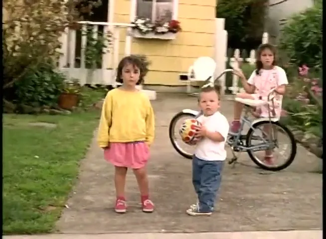 Three children, two girls and a boy, stand on a suburban driveway, looking at the camera as a car pulls away.