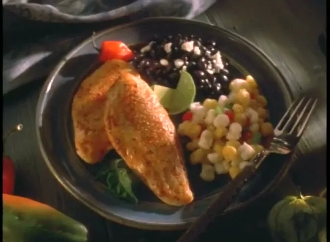 A close-up shot of a plate of food, featuring two chicken breasts, black beans, corn salad, and a lime wedge, with a fork on the side.