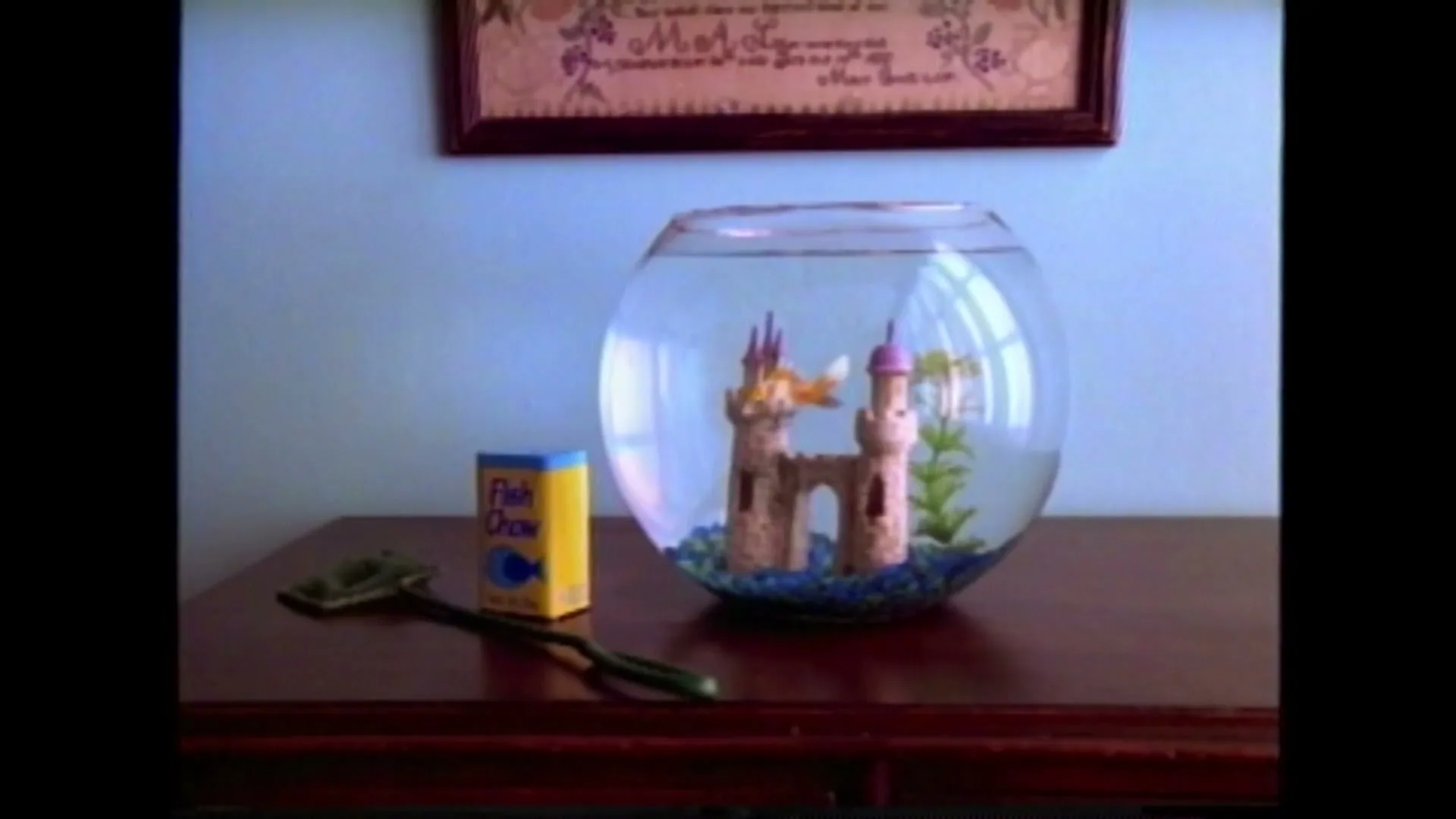 A fishbowl with a castle decoration and a goldfish is on a wooden table next to a box of fish food and a net, in a room with a light blue wall.