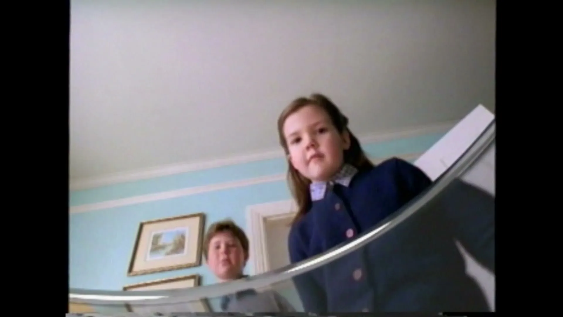 Two children look down into a fishbowl, their faces reflected in the curved glass, in a brightly lit room.