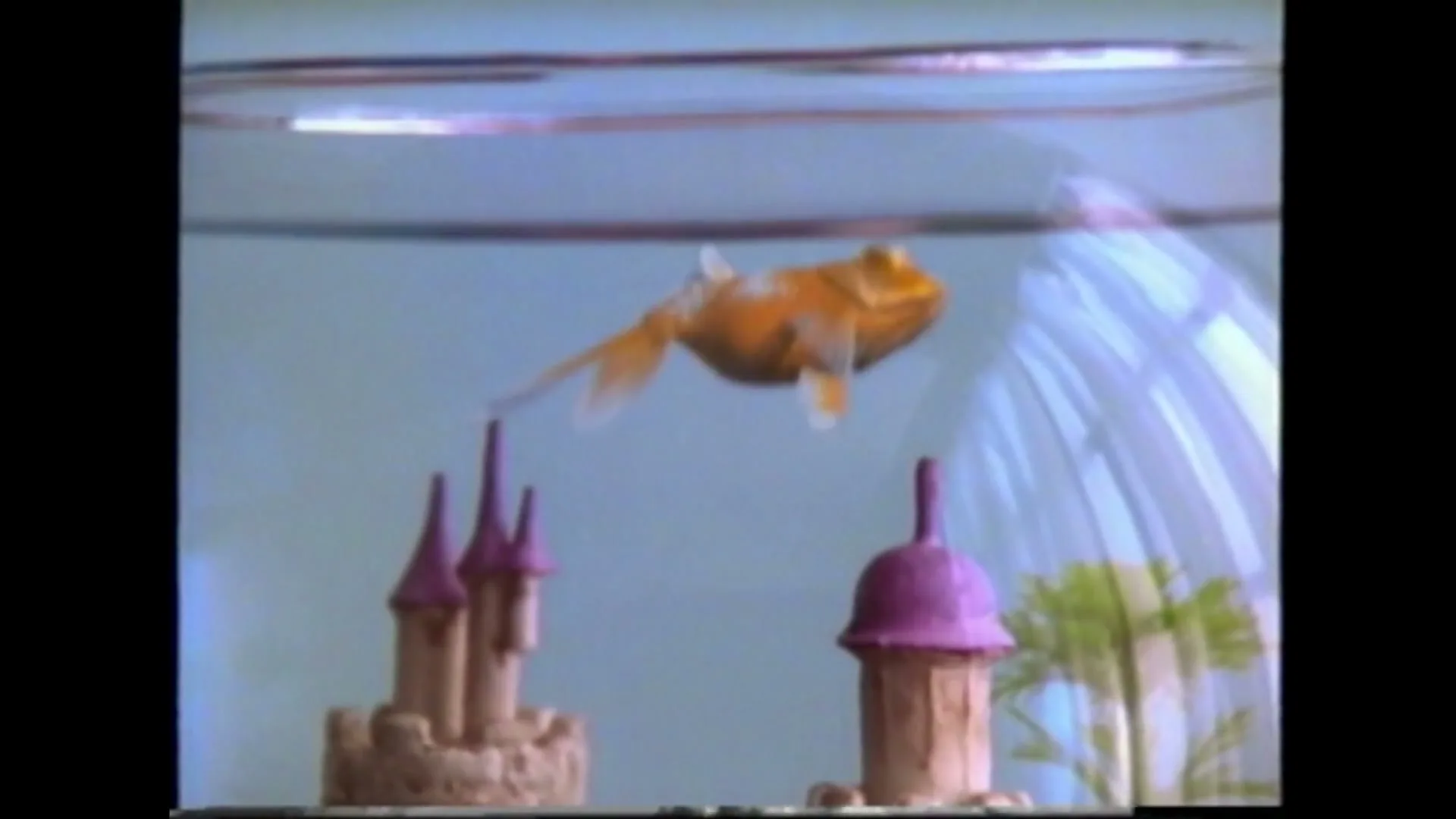 A goldfish with large eyes swims in a fishbowl with a small castle and a purple tower, in a bright room.