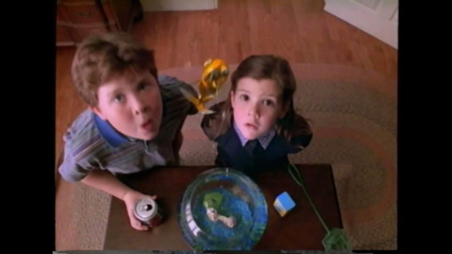 A boy and a girl look down at a goldfish in a bowl on a coffee table, then look up at the camera with surprised expressions in a living room.