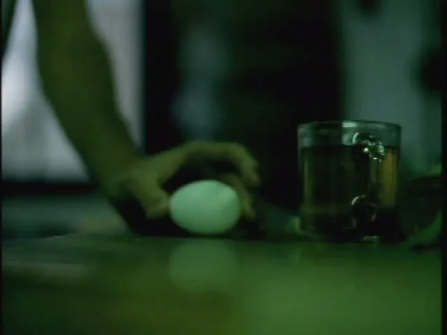 A person's arm and hand are visible as they place a white egg on a wooden table next to a glass of water in a dimly lit room.