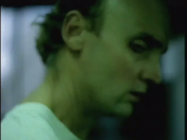 A man with short hair and a white t-shirt looks down and then up, with a green tint to the lighting.