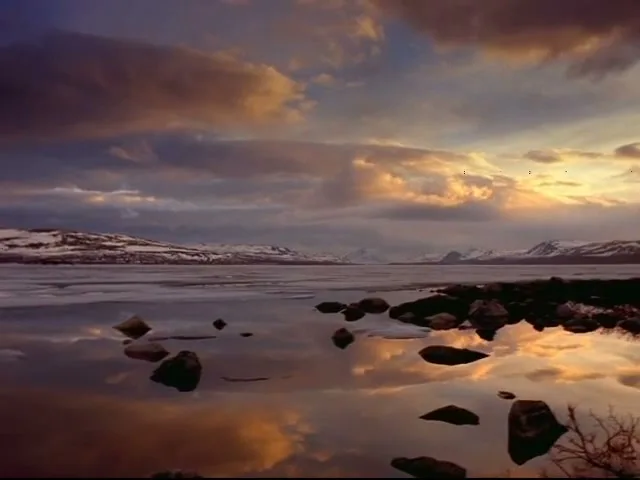 A wide shot of a serene, snow-covered landscape with a calm lake reflecting the colorful sky at dusk, featuring scattered rocks in the foreground.