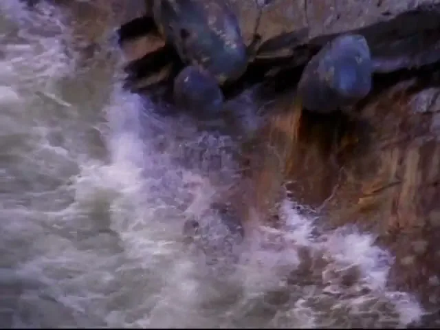 A close-up shot shows several large, dark rocks tumbling down a rocky cliff face into a rushing river, creating splashes.