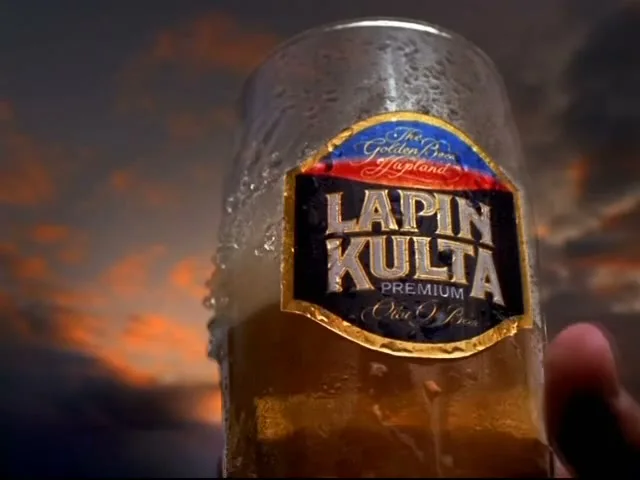 A hand holds a cold glass of Lapin Kulta beer, with condensation and foam, against a dramatic sunset sky over mountains.