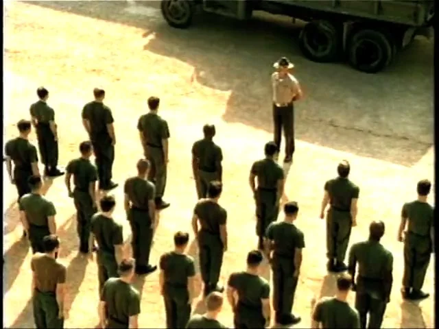 A drill sergeant stands in front of a group of recruits in military uniforms, who are standing at attention in a dusty outdoor area.