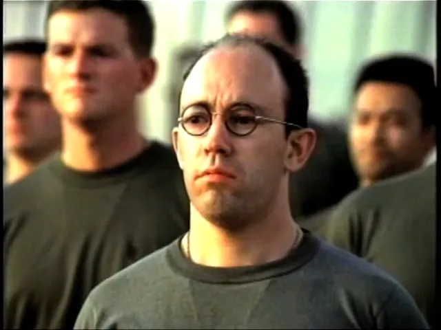A bald man with round glasses and a serious expression stands in a line of soldiers in olive green shirts during the day.