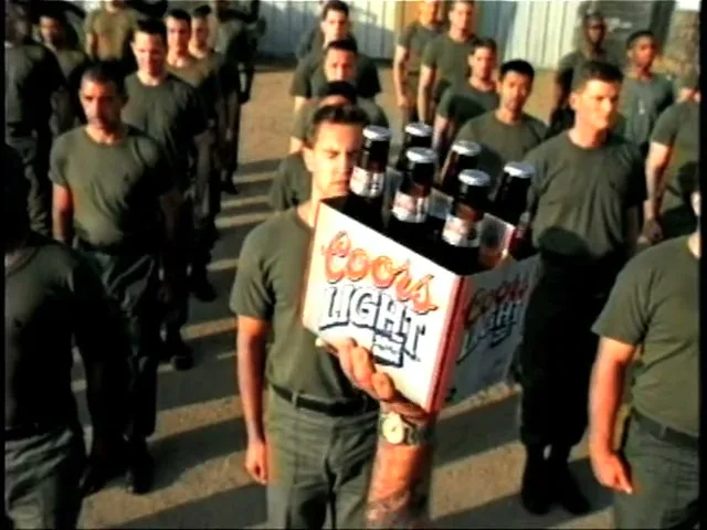 A male soldier in a green uniform stands at attention, holding a 6-pack of Coors Light beer, surrounded by other soldiers in a military formation outdoors.