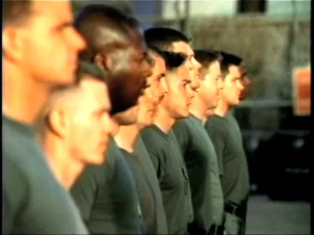 A line of men in military-style uniforms stand at attention, looking straight ahead in an outdoor setting.